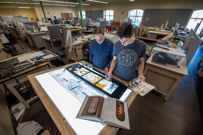 Professor Stephen Hartley stands at a light table with a student examining his luminary.