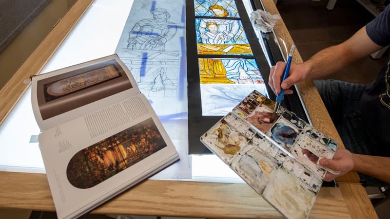 Students painting a luminaire on a lightbox with paintbrush and palette in hand.