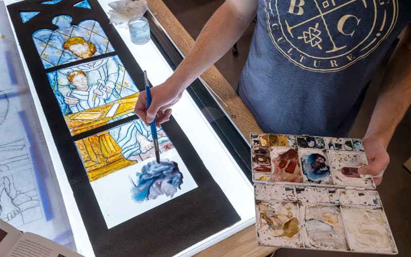 A students paints a luminaire — a mockup of the window painted onto clear acetate and framed, so that light can shine through it 