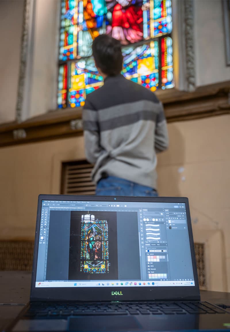 A laptop with an image of a stained glass window is in the foregound, with a male student standing at a stained glass window in the background.