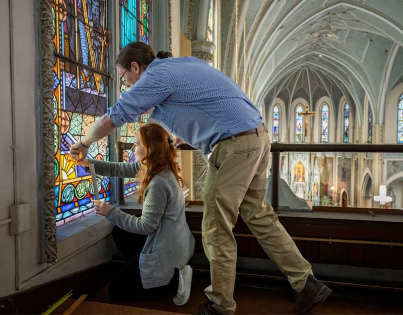 A female student kneels at the base of a stained glass window taking measurements while a male professor stands above her, helping.