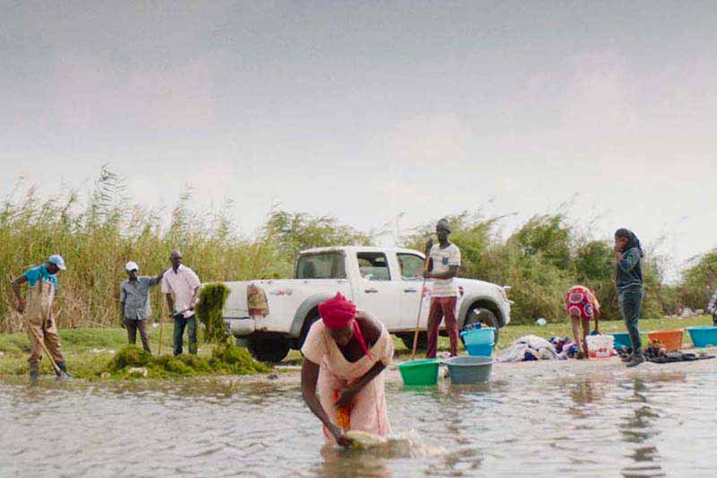 One woman in the foreground stands in the middle of a river and bends over fishing for something in the water. In the background, several people stand around a truck with buckets and shovels.