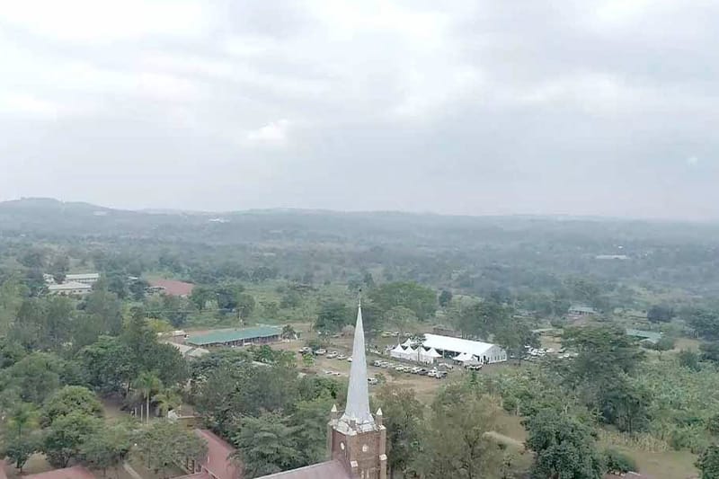 An aerial view of an area of Uganda. A large white tent and several cars parked, surrounded by trees.