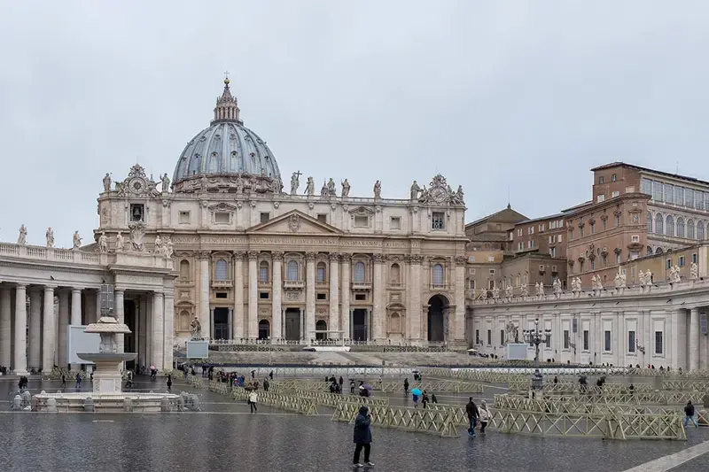 St. Peter's Basilica in Vatican City on a cloudy day.  People walk through the large, wet plaza, partially cordoned off with temporary barriers.