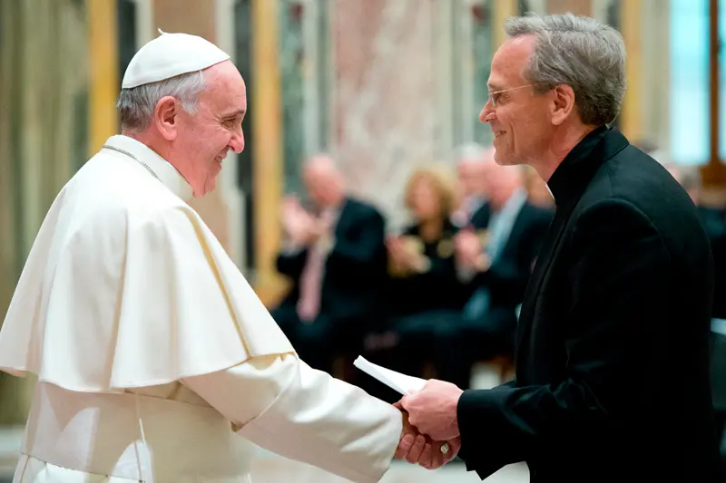 Pope Francis, wearing white papal vestments, smiles and shakes hands with a man in a dark suit and clerical collar, while also handing him a small white booklet.  A blurred audience is visible in the background.