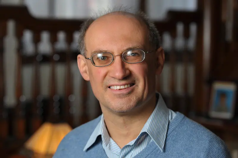 Headshot of a man with glasses, wearing a light blue sweater over a light blue and white striped collared shirt, smiling at the camera.  A blurry background shows wooden furniture and a lamp.