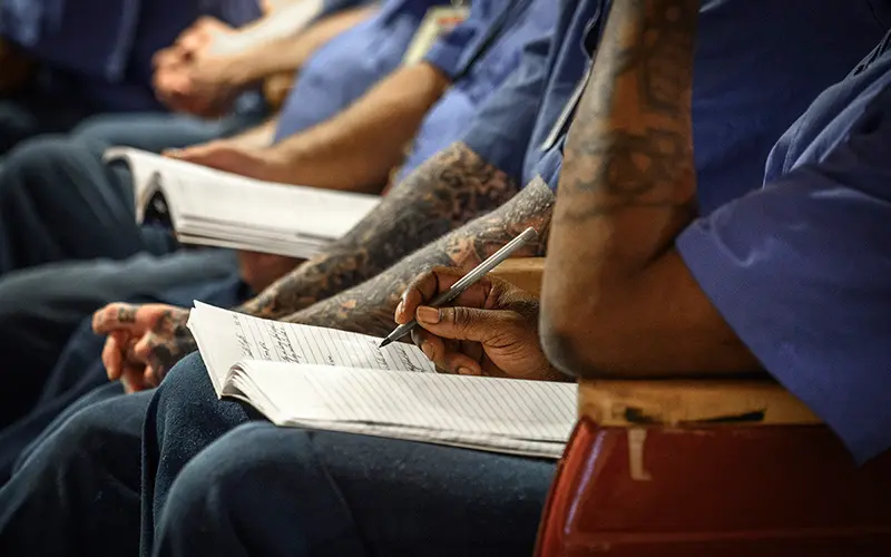lose-up of several individuals wearing blue attire, likely incarcerated individuals, sitting in rows. Focus is on the hands of one person writing in a notebook with a pen. Their arms are adorned with tattoos.  Others in the row also appear to be taking notes in a classroom or meeting setting.