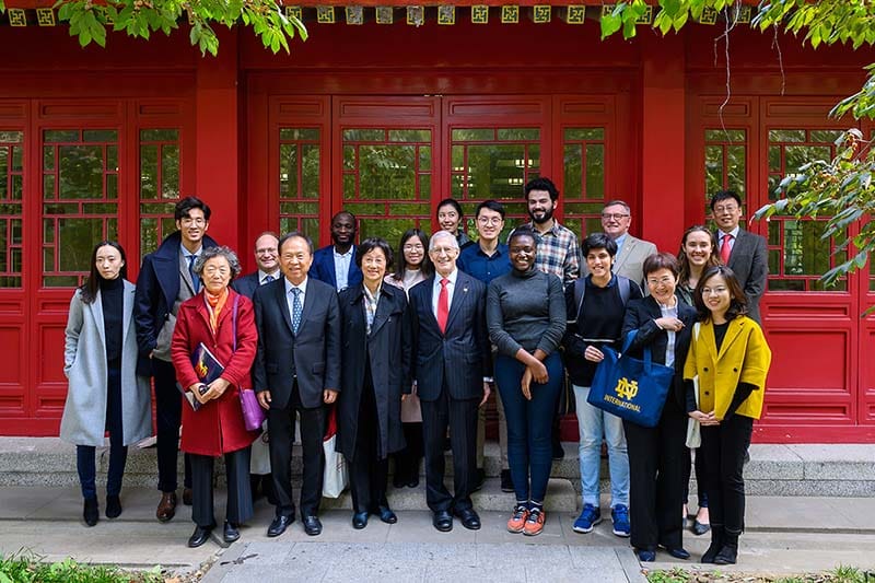 A group of Notre Dame delegation and students and staff at Peking University stand together in from of a red building.