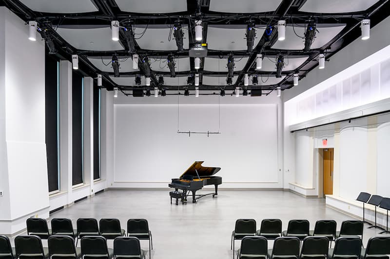 A modern white open space containing black chairs lined in neat rows on the left and right of the aisle. A sleek black baby grand piano stands empty at the front of the room.