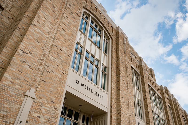 An impressive looking brick building towers above the photographer. Above the door, a sign reads 'O'Neill Hall.' The tall windows reflect blue skies and white clouds.