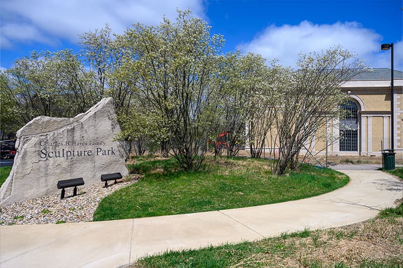 A large boulder with 'Sculpture Park' inscribed sits forward and left of the Raclin Murphy Museum of Art. A large green bush surrounds the boulder and a pathway leads visitors from the park to the museum.