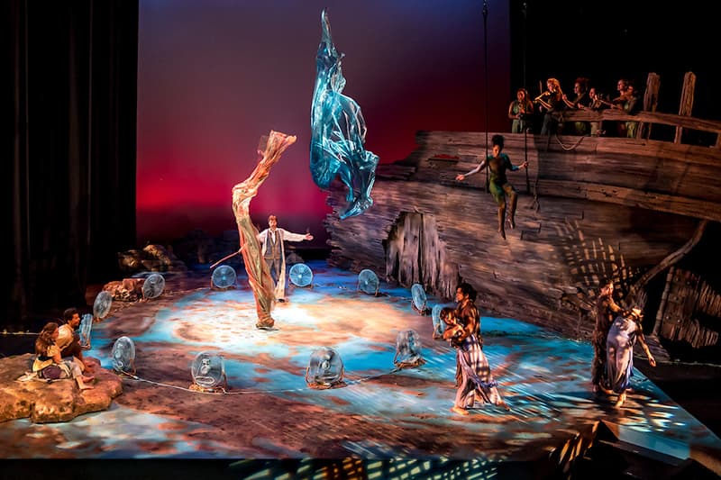 Actors perform on a stage lit in blue and gold. An aerialist swings about the stage as actors throw colorful fabric into the air.