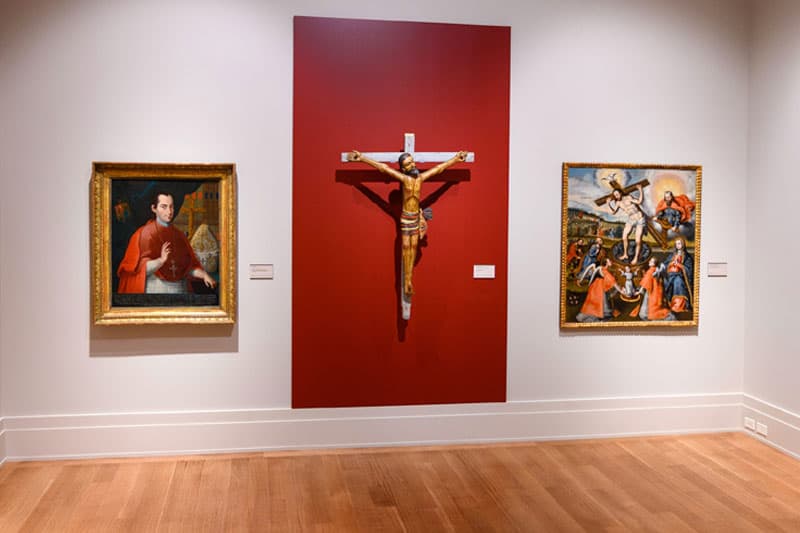 A cross on a red wall flanked by two Christian paintings on white walls.