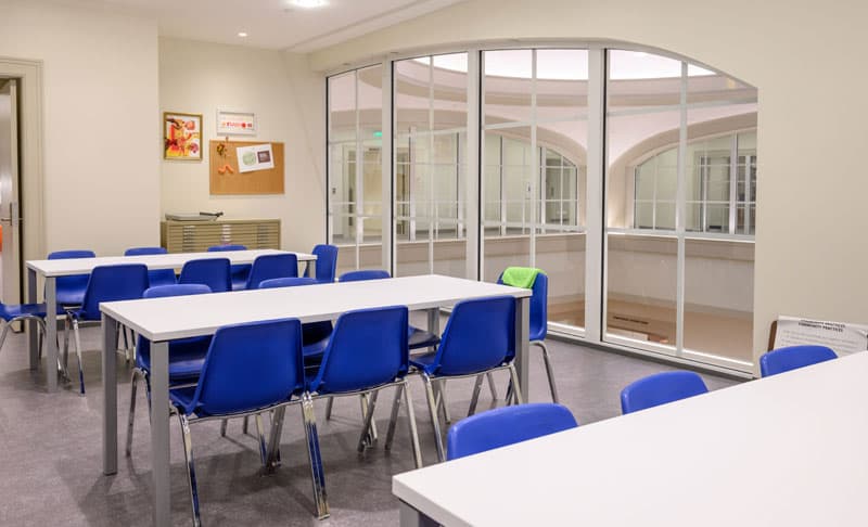 A bright open space with a large window, three long white tables, and blue chairs.