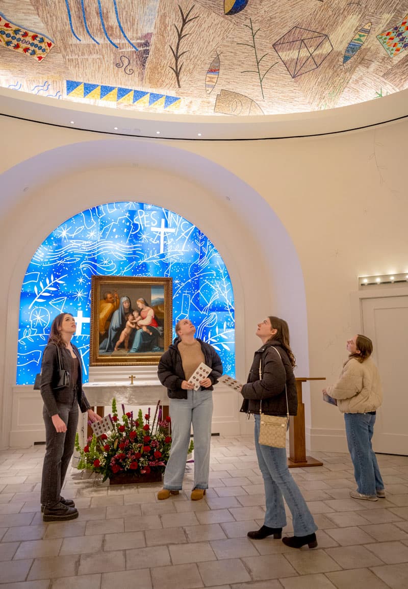 Four students stand in the middle of the Queen of Families Chapel looking up at the decorated ceiling.