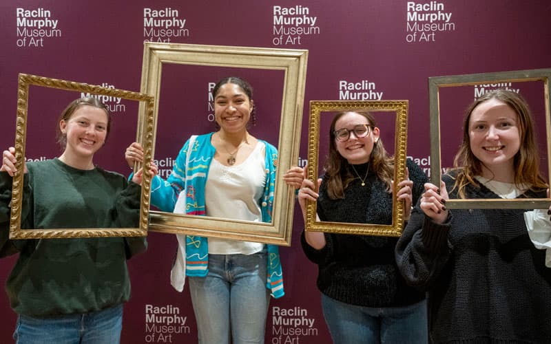Four students stand against a purple backdrop holding empty picture frames around their faces.