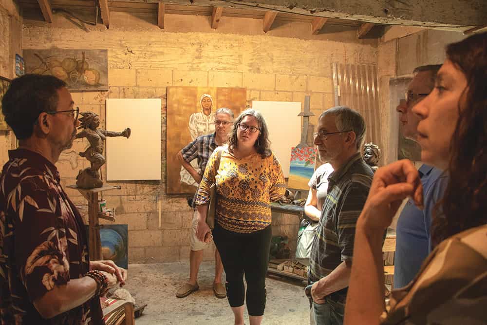 A group of people stand around listening to another talk about the canvas paintings in the room.