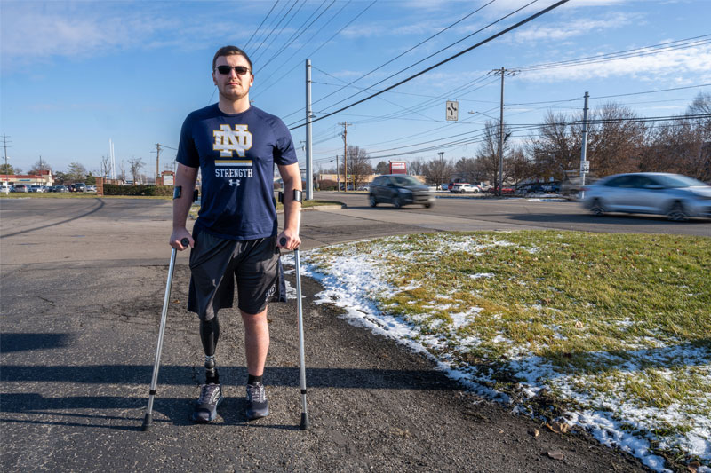 Andrew stands at the spot where his accident happened. He's wearing his prosthetic leg and using crutches.