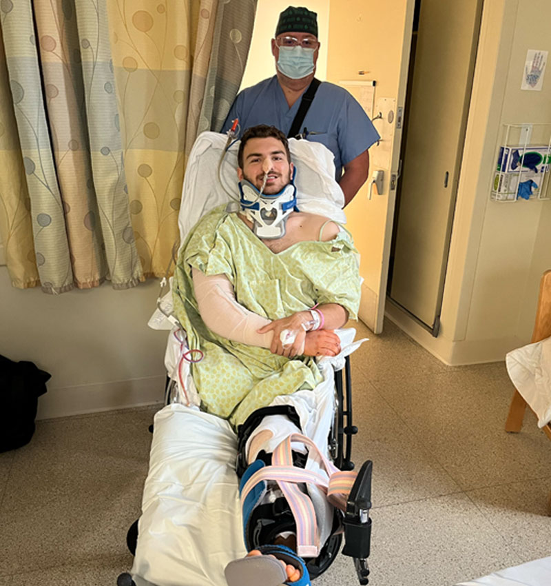 Andrew is being pushed in his wheelchair by his nurse. He has on a neckbrace, tubes running from his nose, a broken arm, and you can see where his leg is missing.