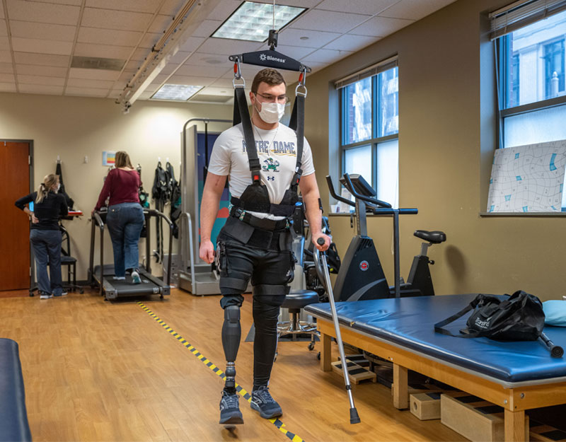 Andrew is strapped into a harness that is suspended by the ceiling, helping him learn to walk with the prosthetic.