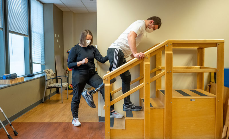 Andrew is doing physical therapy by doing exercises on a set of stairs. His physical therapist stands behind to assist.