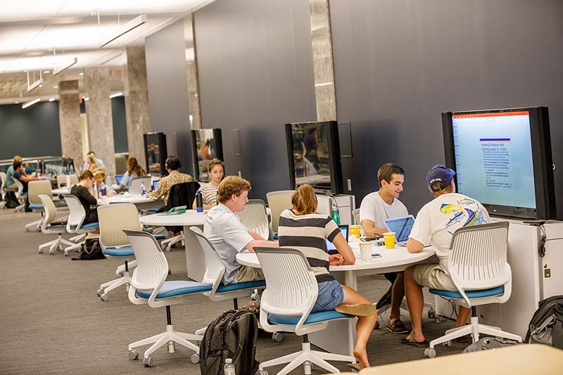 A group of students sit at a table together, looking at a screen mounted above the table with work on it. Other similar tables are in the background.