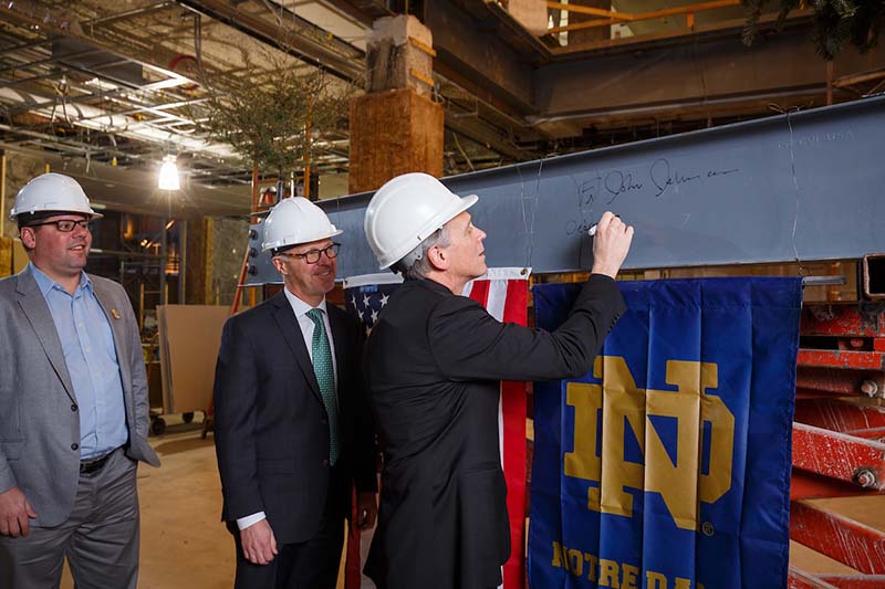 Father Jenkins, wearing a hard hat, signs a steel beam with a ND monogram flag hanging from it while colleagues look on.