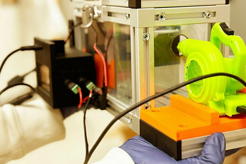 Bright green, plastic impeller attached to an orange mount inside a clear and metal experimental box. A gloved hand holds the device.  Wires and a separate black box are also visible.