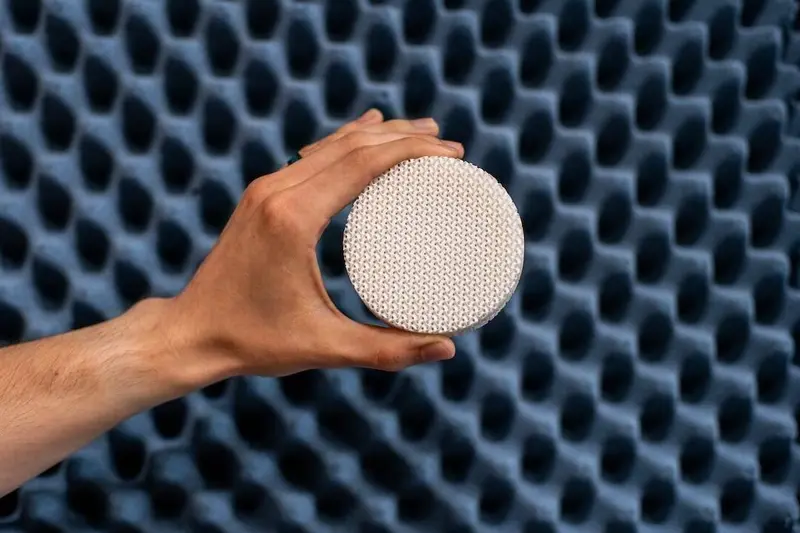 A hand holds a small, circular, white object with a textured, woven pattern. The backdrop is a dark blue surface with a repeating, geometric pattern of concave shapes.