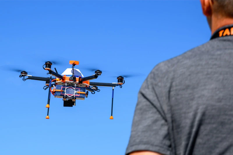 An orange and white drone with black propellers flies in a clear blue sky.  A person wearing a gray shirt observes the drone.