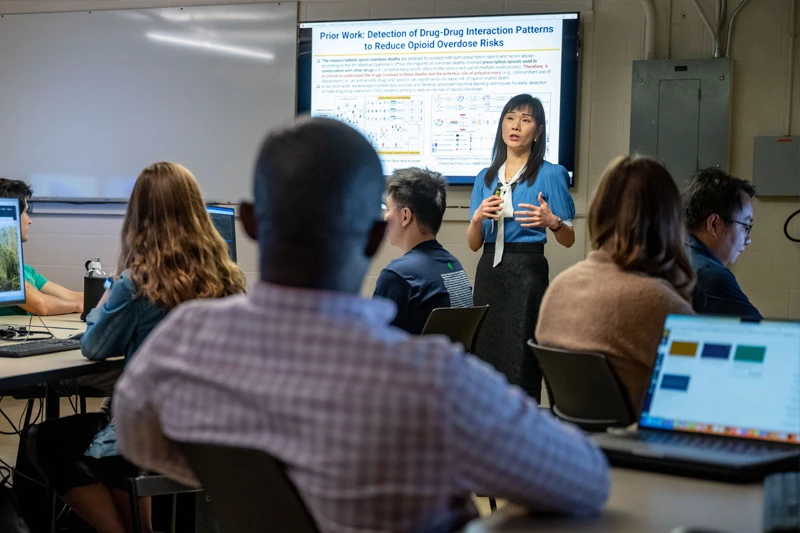 A woman in a blue shirt and black skirt gestures as she presents to a classroom of students.
