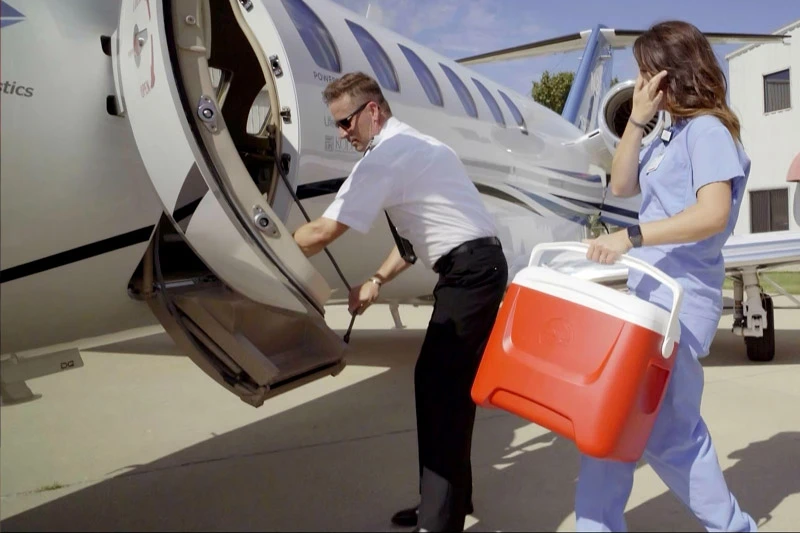 Pilot lowers the stairs of a private jet as a medical professional in blue scrubs carries a red and white cooler from the plane.