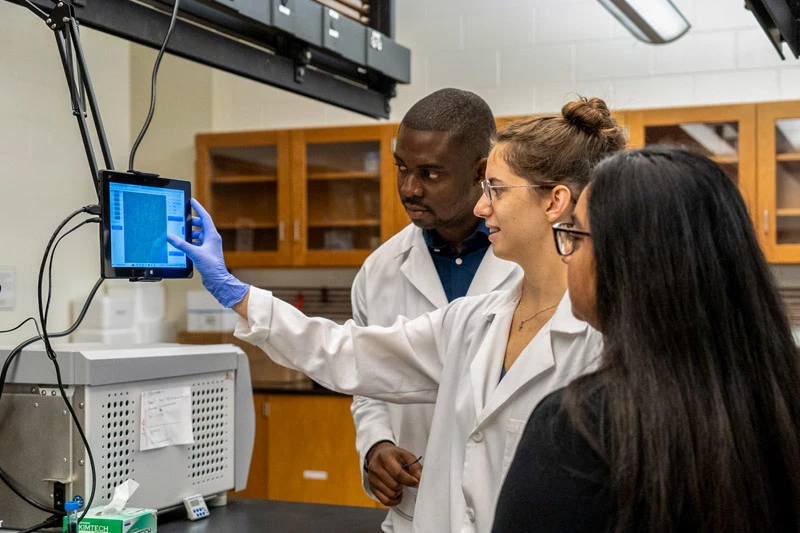 Three scientists in white lab coats examine data on a tablet screen in a University of Notre Dame laboratory.  The scientist on the left points at the screen with a gloved hand while her colleagues observe. A laboratory device is visible in the bottom left corner.