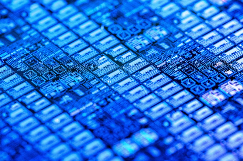 Close-up of a microchip bathed in blue light. The intricate circuitry and repetitive square units create a visually striking pattern.
