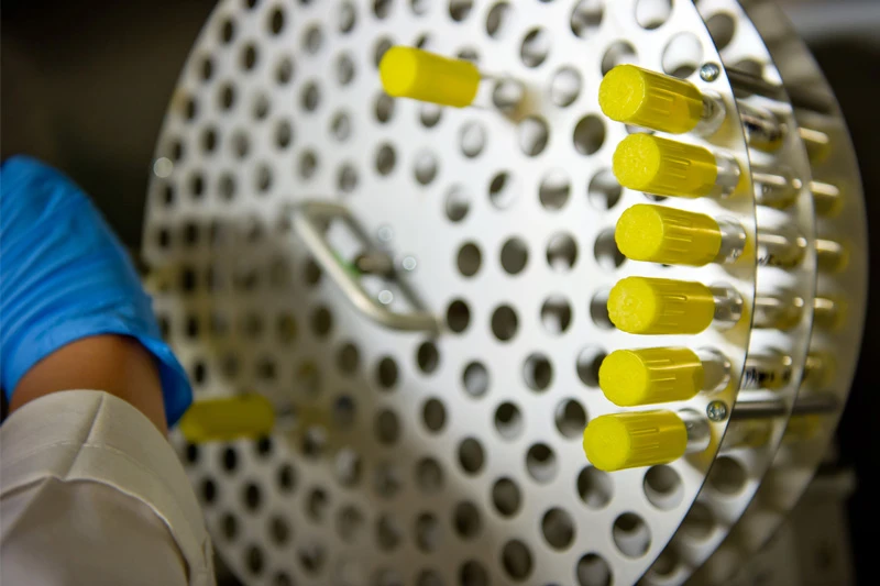 Close-up of a researcher's gloved hand reaching towards a circular metal rack containing several vials filled with a bright yellow substance. The rack has numerous small holes, and the vials are secured within a designated section.