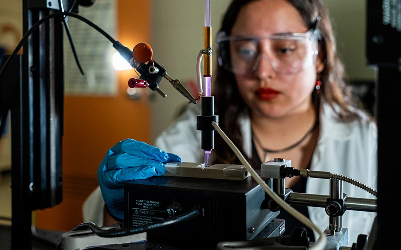 A student wearing safety glasses and blue gloves uses a scientific instrument emitting a purple plasma beam.