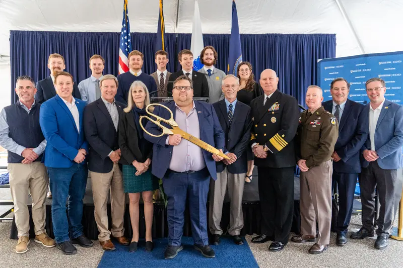 A group of men and women pose for a photo. A person in the center holds large, gold ceremonial scissors, suggesting a ribbon-cutting ceremony. They stand on a blue carpet in front of a navy backdrop with flags and a banner that says "Naval Surface Warfare Center."