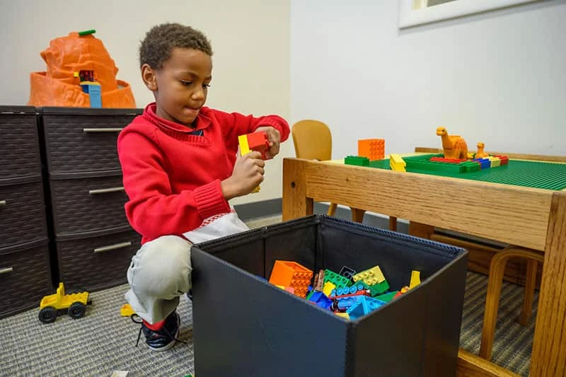 A black boy in red sweatshirt leans over a bin of colorful plastic blocks.