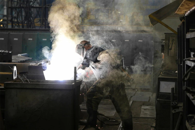 A welder wearing a protective mask and dark clothing works on a metal container, surrounded by bright sparks and smoke in an industrial setting.