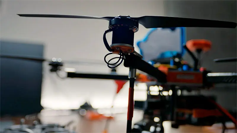 Close-up of a drone's rotor and arm. sThe black rotor blades are slightly blurred, suggesting motion. Black and orange components of the drone are visible in the background.
