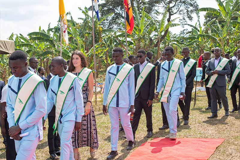 Recent graduates wearing sashes walk down a path with a red carpet on the ground.