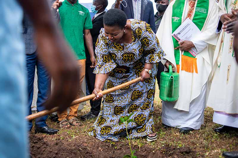 A woman wearing colorful traditional clothing holds onto a gardening hoe and pushes dirt over a newly planted tree.