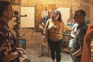 A group of people stand around listening to another talk about the canvas paintings in the room.