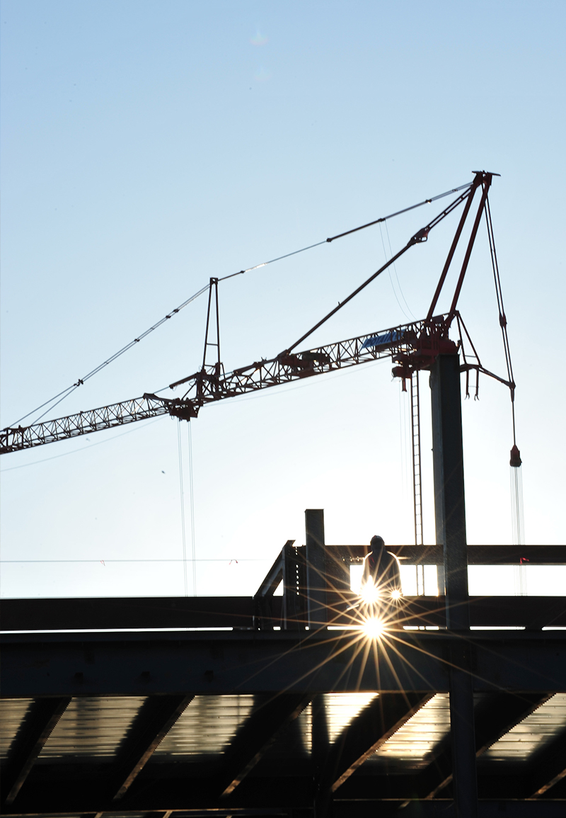 A man in a hardhat stands atop metal beams. A large crane is behind him. Sunlight filters through and creates two bursts of light next to him. 