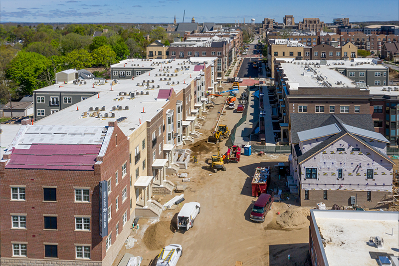 An aerial view of Eddy Street during phase two of the project. The road is dirt with CAT construction trucks and vans parked along it. Buildings are in various stages of build, some have brick siding while others are still waiting for roofs.