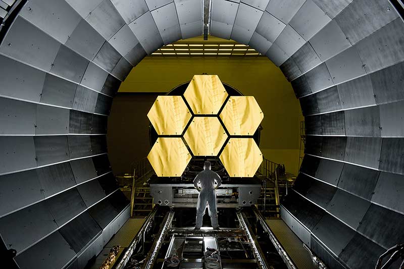A person with their back to the camera stands in front of a part of Webb's telescope featuring 6 golden hexagonsf, wearing a cleanroom suit stands with their legs shoulder-length apart, their hands on their hips.