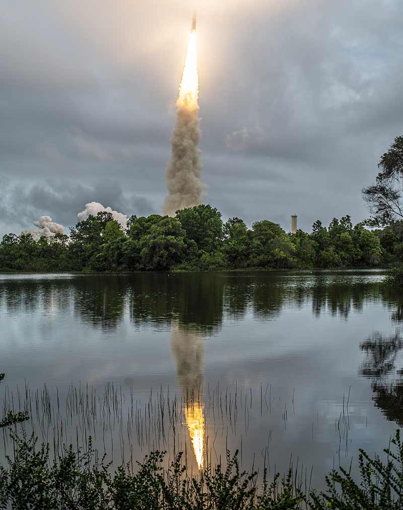 Smoke billows out of a rocket that launches from a distance and is rerflected off of a river.