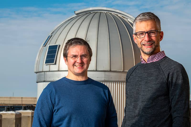 Two male professors stand side by side outside in front of an observatory dome.