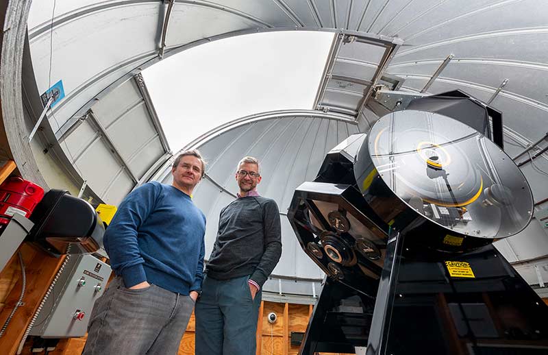 Two male professors stand side by side inside of an observatory dome looking down towards the camera.