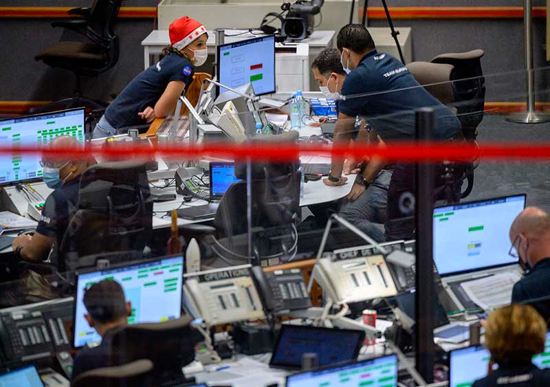 A female at a desk, wearing a red Santa hat, leans forward to talk to two  males looking at a computer. They talk amongst themselves. Others sit at computers in the foreground which is out of focus.